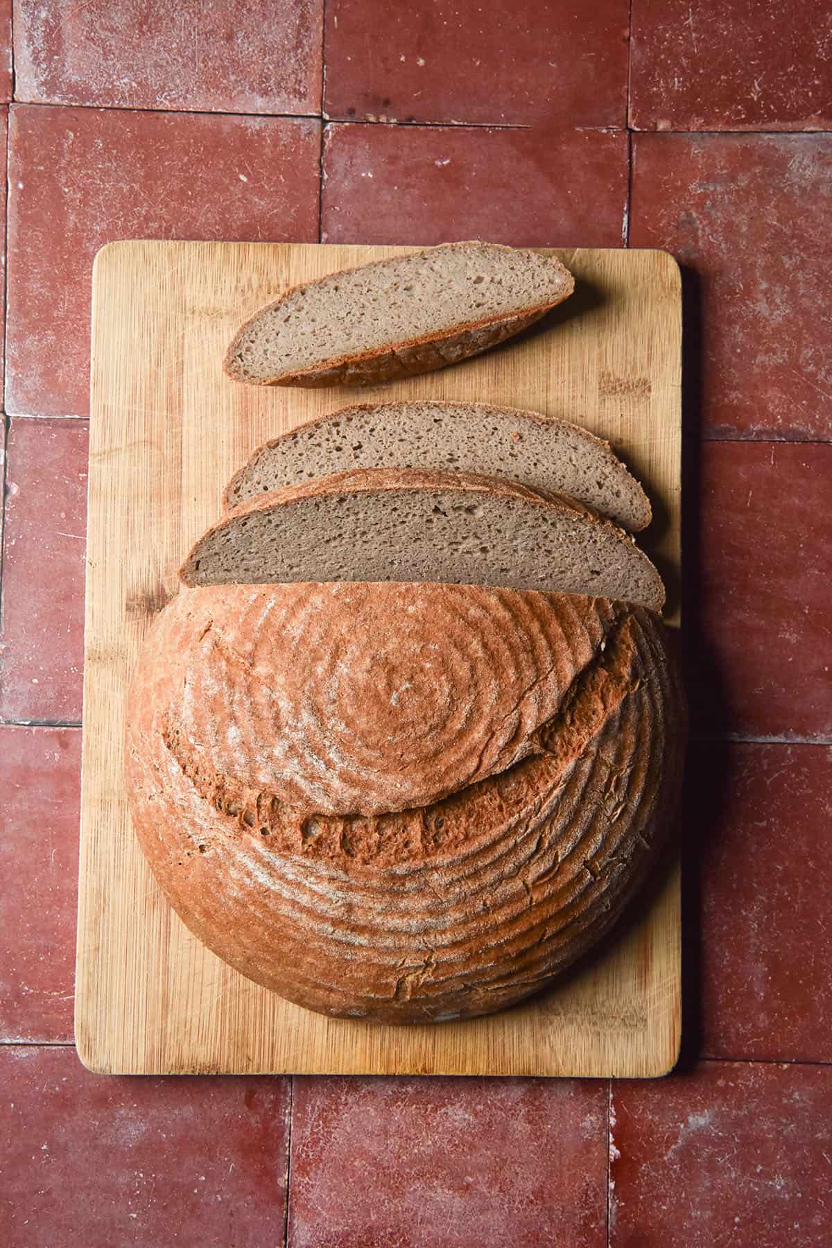 An aerial image of a buckwheat boule on a chopping board that has the top three slices cut and casually arranged on the board. The board sits atop a terracotta tile backdrop