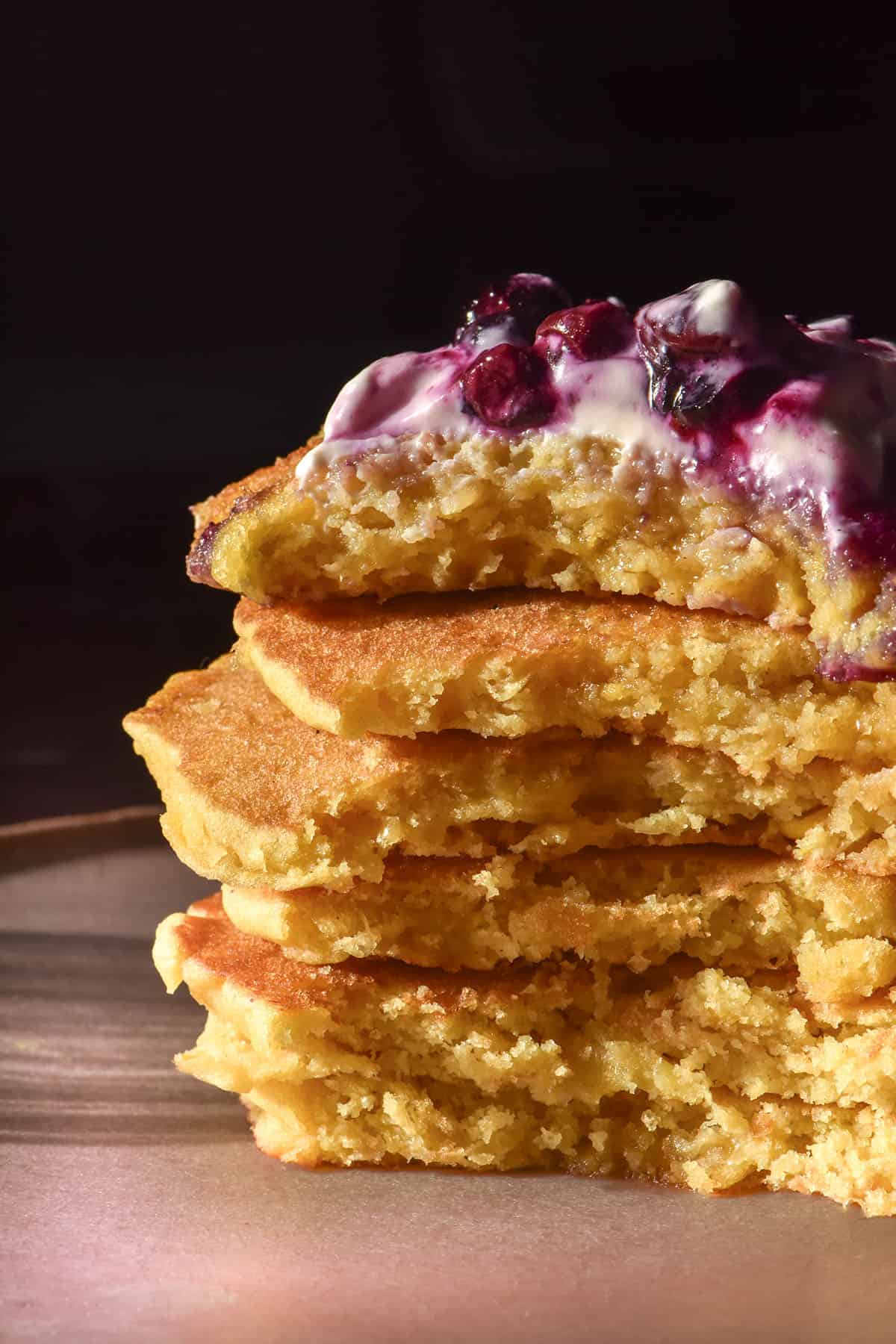 A moody side on image of a stack of gluten free cornmeal pancakes topped with yoghurt and a blueberry coulis. The stack sits on a pale pink ceramic plate against a dark backdrop.