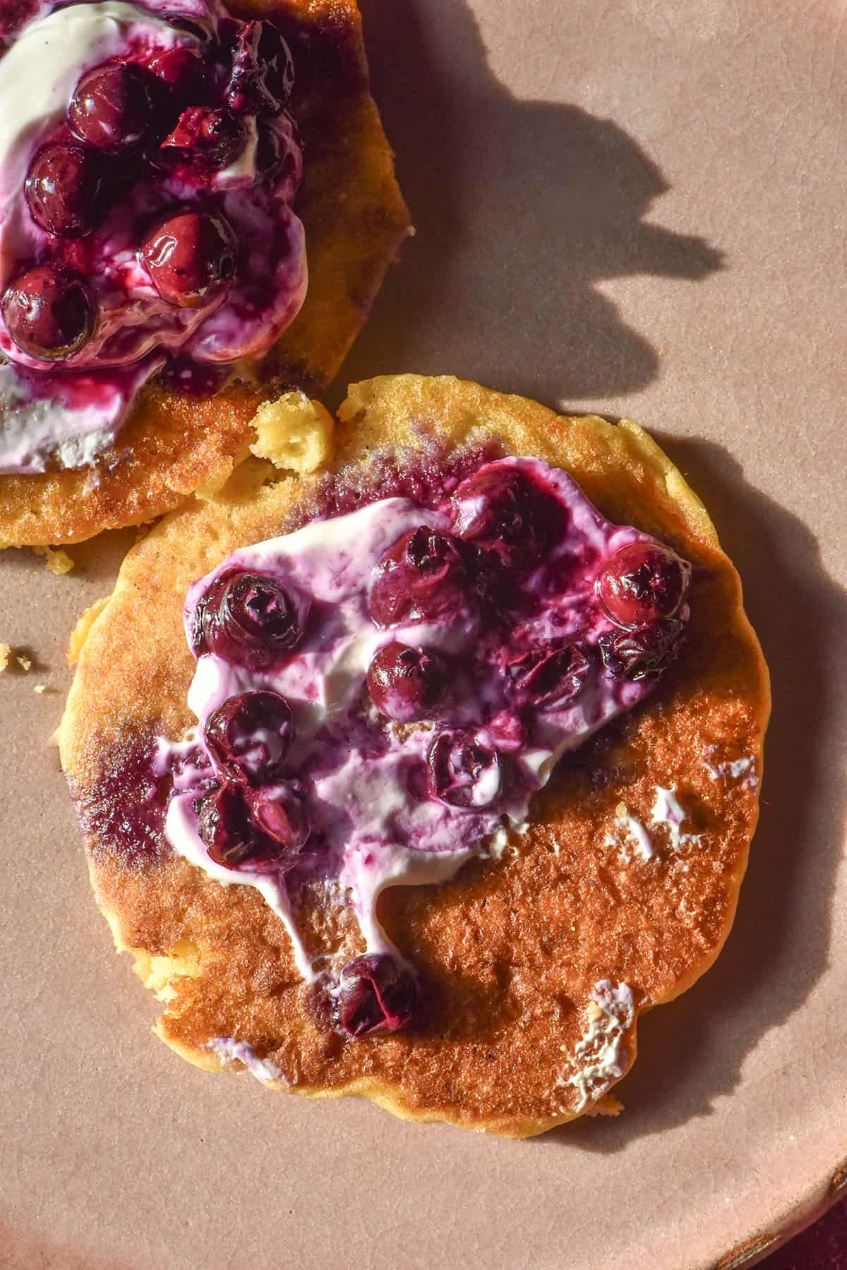 An aerial image of cornmeal pancakes topped with yoghurt and blueberry coulis on a pale pink ceramic plate