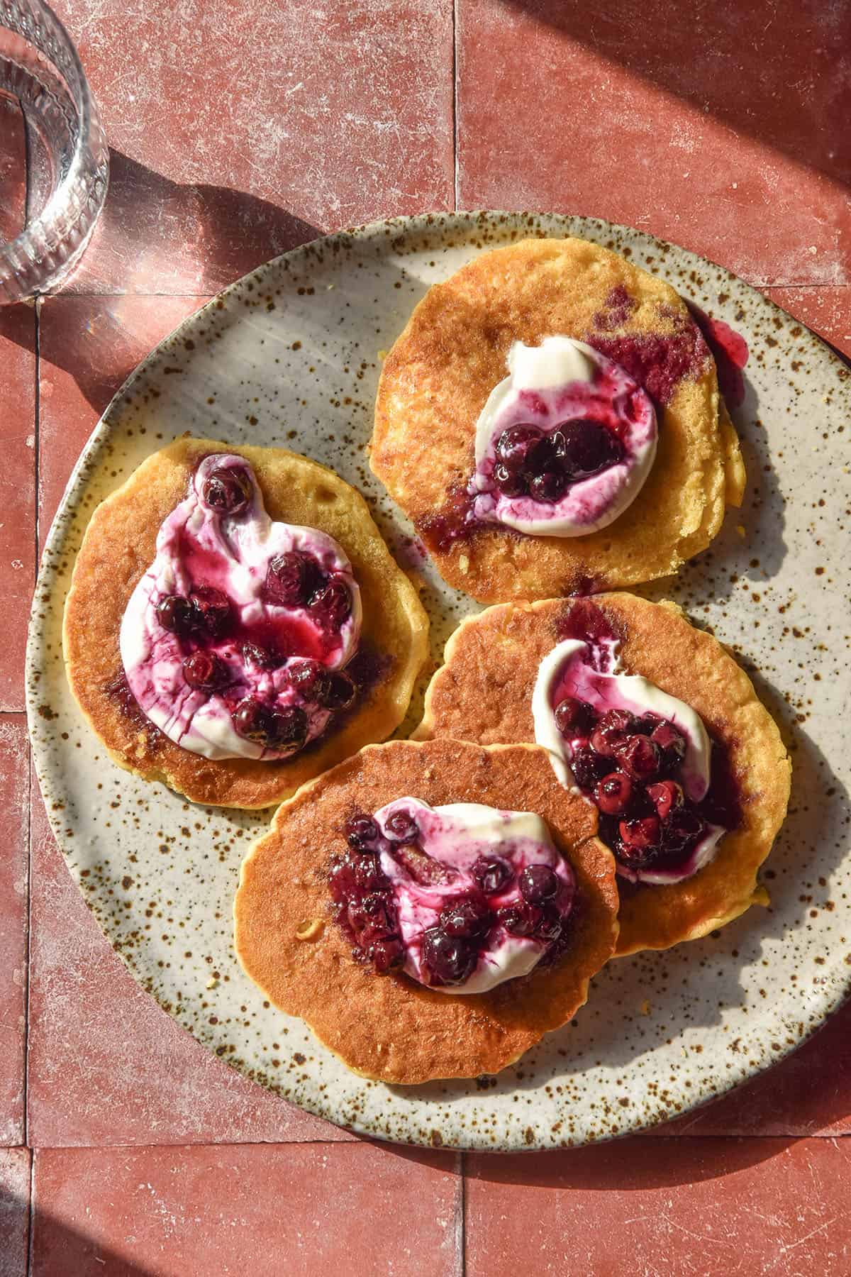 An aerial image of a white speckled ceramic plate atop a terracotta tile backdrop topped with gluten free cornmeal pancakes. The pancakes are topped with yoghurt and blueberry coulis and a sunlit glass of water sits in the top left of the image