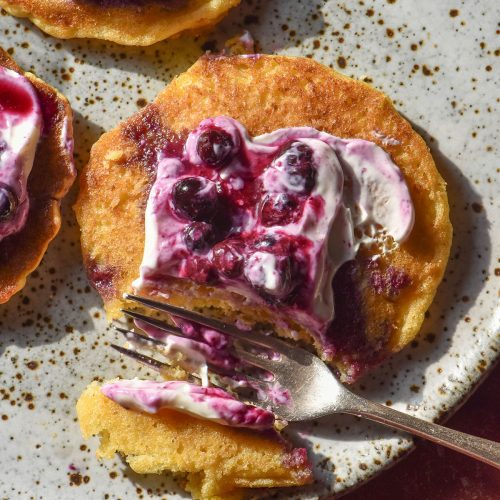 An aerial image of gluten free cornmeal pancakes topped with blueberry coulis and yoghurt on a white speckled ceramic plate atop a terracotta tile backdrop