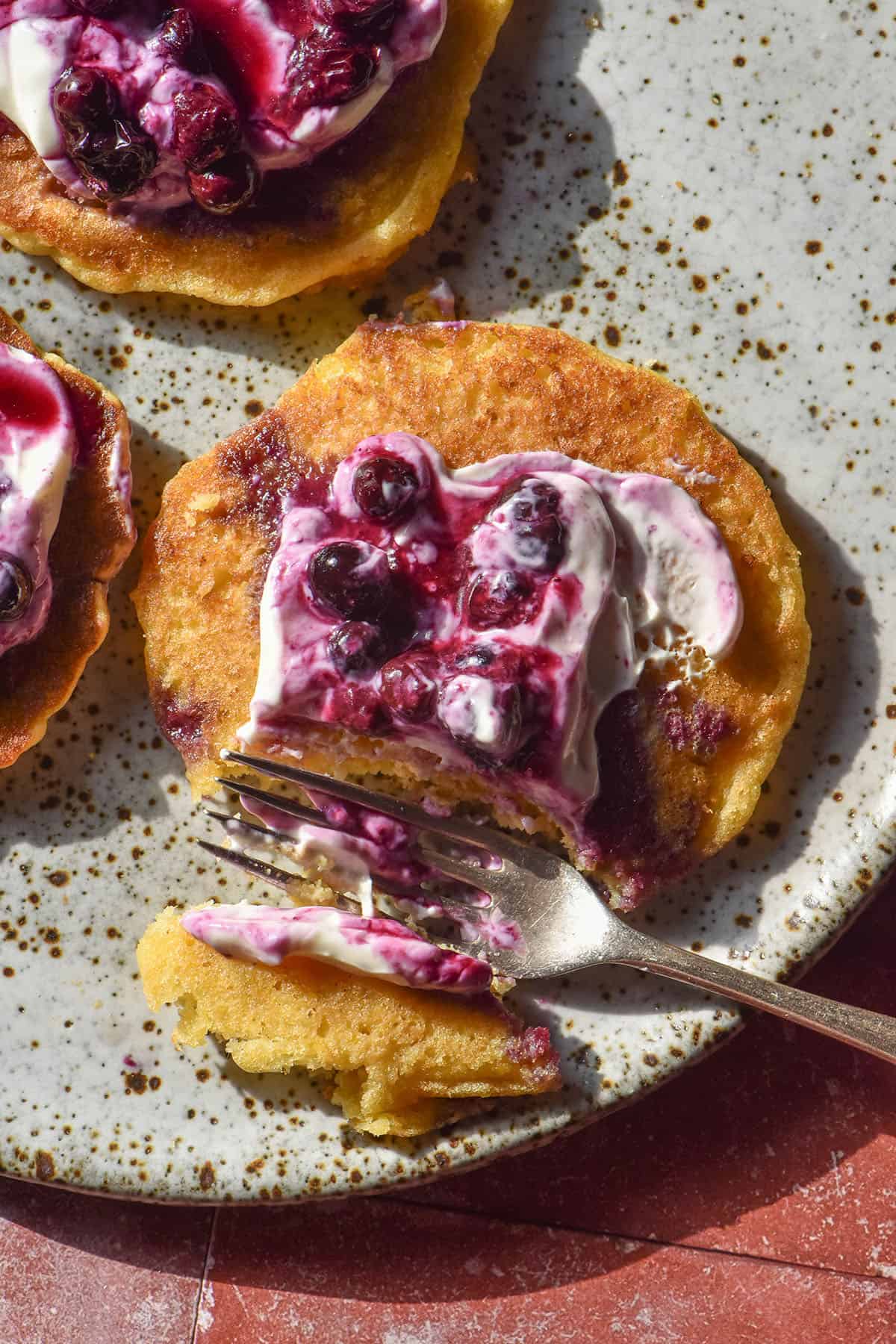 An aerial image of gluten free cornmeal pancakes topped with blueberry coulis and yoghurt on a white speckled ceramic plate atop a terracotta tile backdrop