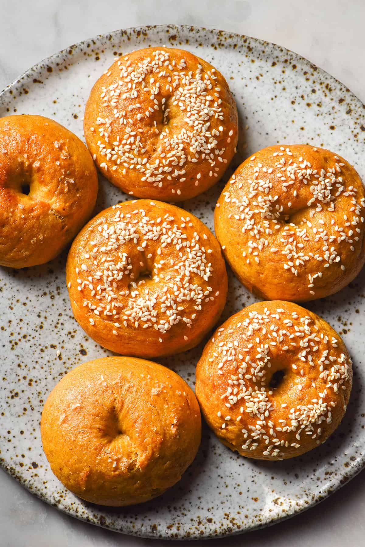 An aerial image of gluten free pumpkin bagels topped with sesame seeds on a white speckled ceramic plate atop a white marble table