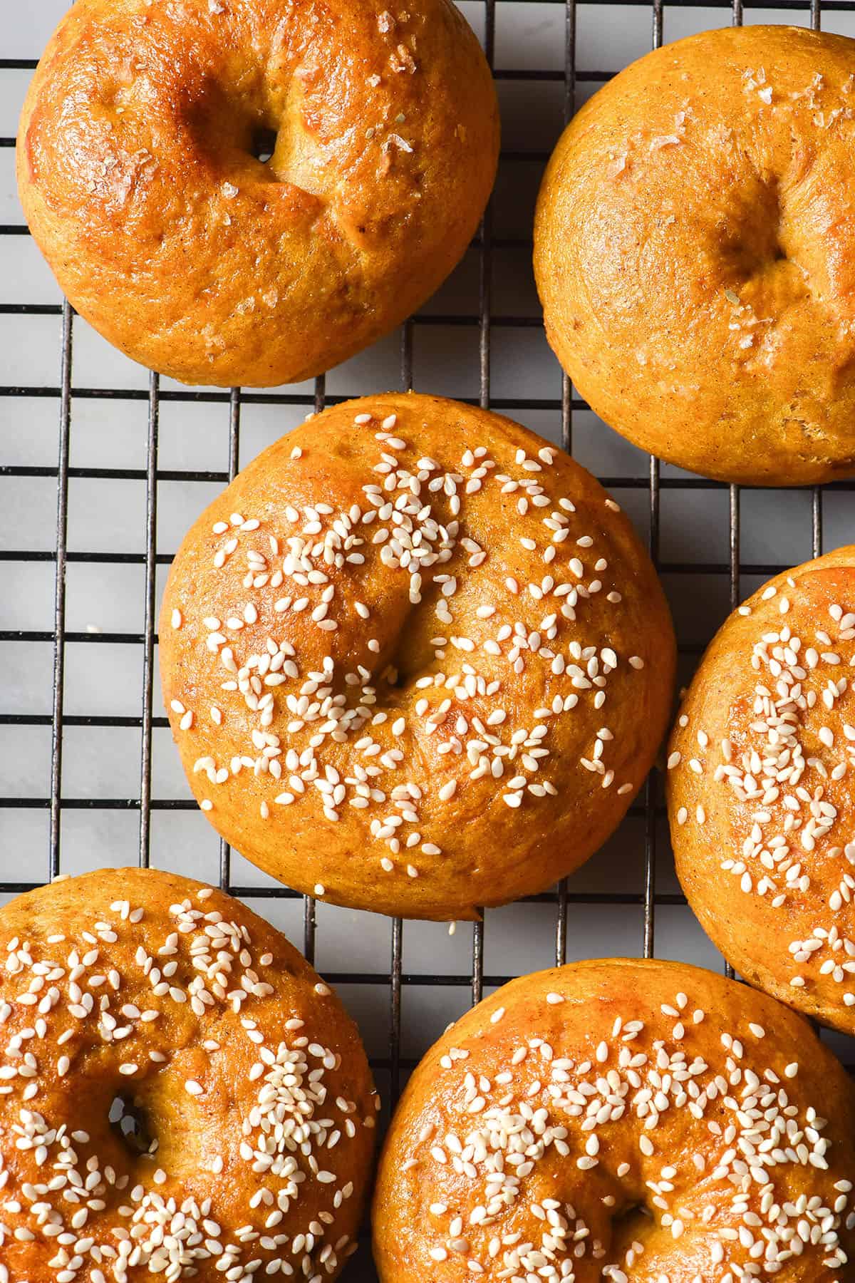 An aerial image of pumpkin bagels topped with sesame seeds on a black wire cooling rack atop a white marble table