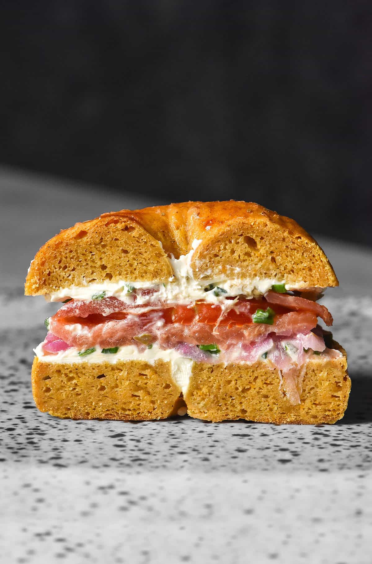 A side on image of a gluten free pumpkin bagel filled with cream cheese, smoked salmon, capers, pickled onion and chives. The bagel has been sliced in half, revealing the fillings. It sits on a white speckled ceramic plate against a dark backdrop