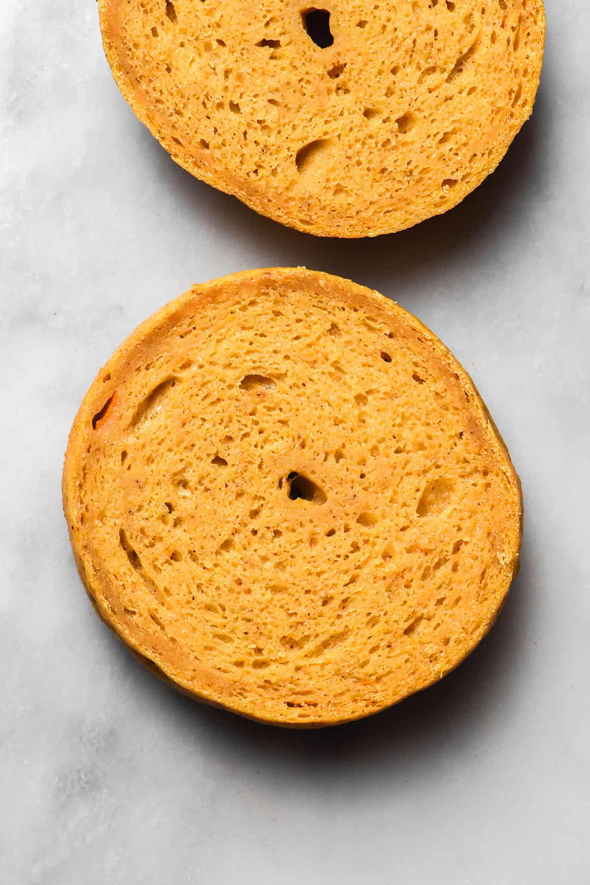 An aerial image of a gluten free pumpkin bagel that has been sliced in half to reveal the inner crumb. Both halves sit on a white marble table.