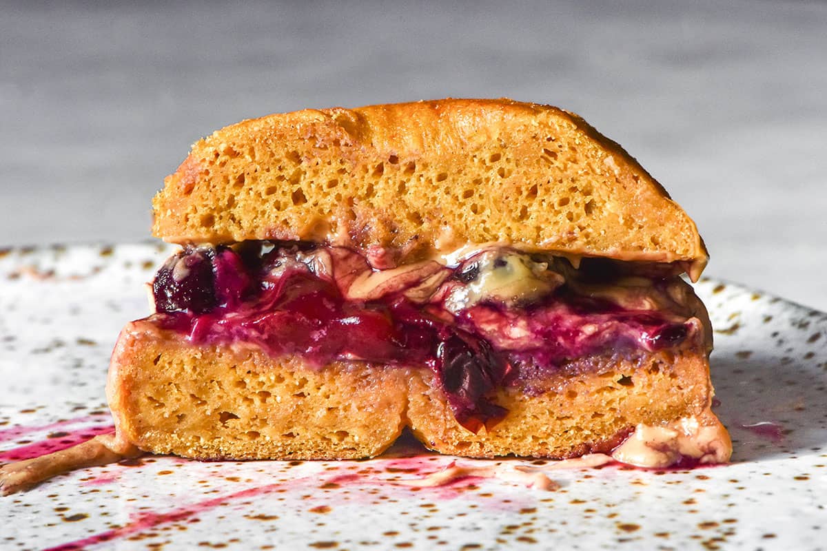 A side on macro image of a gluten free pumpkin bagel filled with blueberry jam and peanut butter. The bagel has been sliced in half to reveal the filling. The bagel half sits on a white speckled ceramic plate against a white marble table