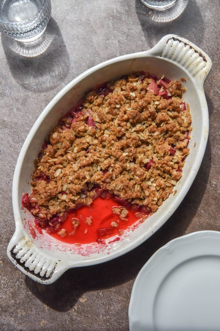 An aerial sunlit image of a white ceramic baking dish filled with gluten free rhubarb crumble. The dish sits atop a grey backdrop in bright sunlight and is surrounded by glasses of water and extra plates