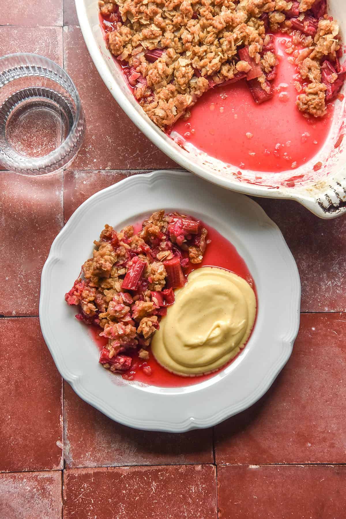 An aerial image of a plate of gluten free rhubarb crumble and lactose free custard atop a terracotta tile backdrop. The dish of crumble sits to the top right of the plate and a sunlit glass of water sits to the top left