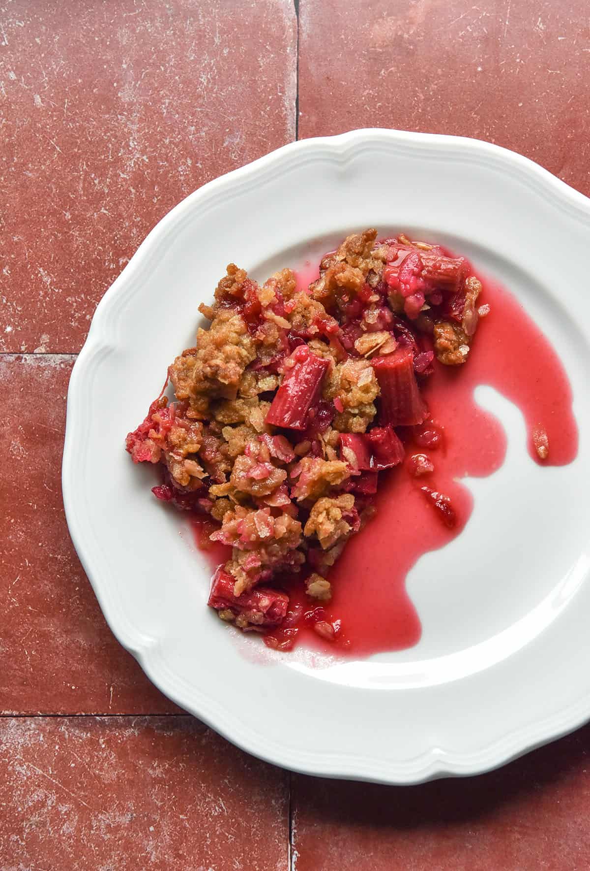 An aerial image of a white plate topped with gluten free rhubarb crumble. The plate sits atop a terracotta tile backdrop