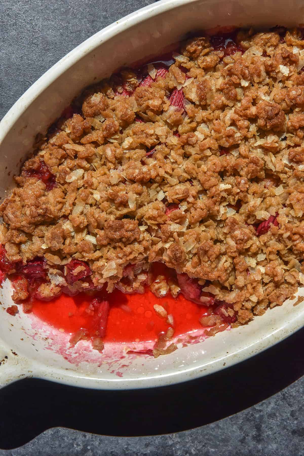 An aerial sunlit image of a gluten free rhubarb crumble in a white oval baking dish. The dish sits atop a dark blue backdrop