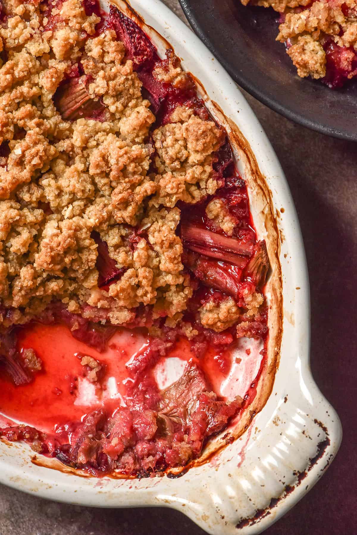 A close up macro image of a white oval dish filled with gluten free rhubarb and strawberry crumble. Some of the crumble has been scooped out, revealing the jammy fruit underneath. 