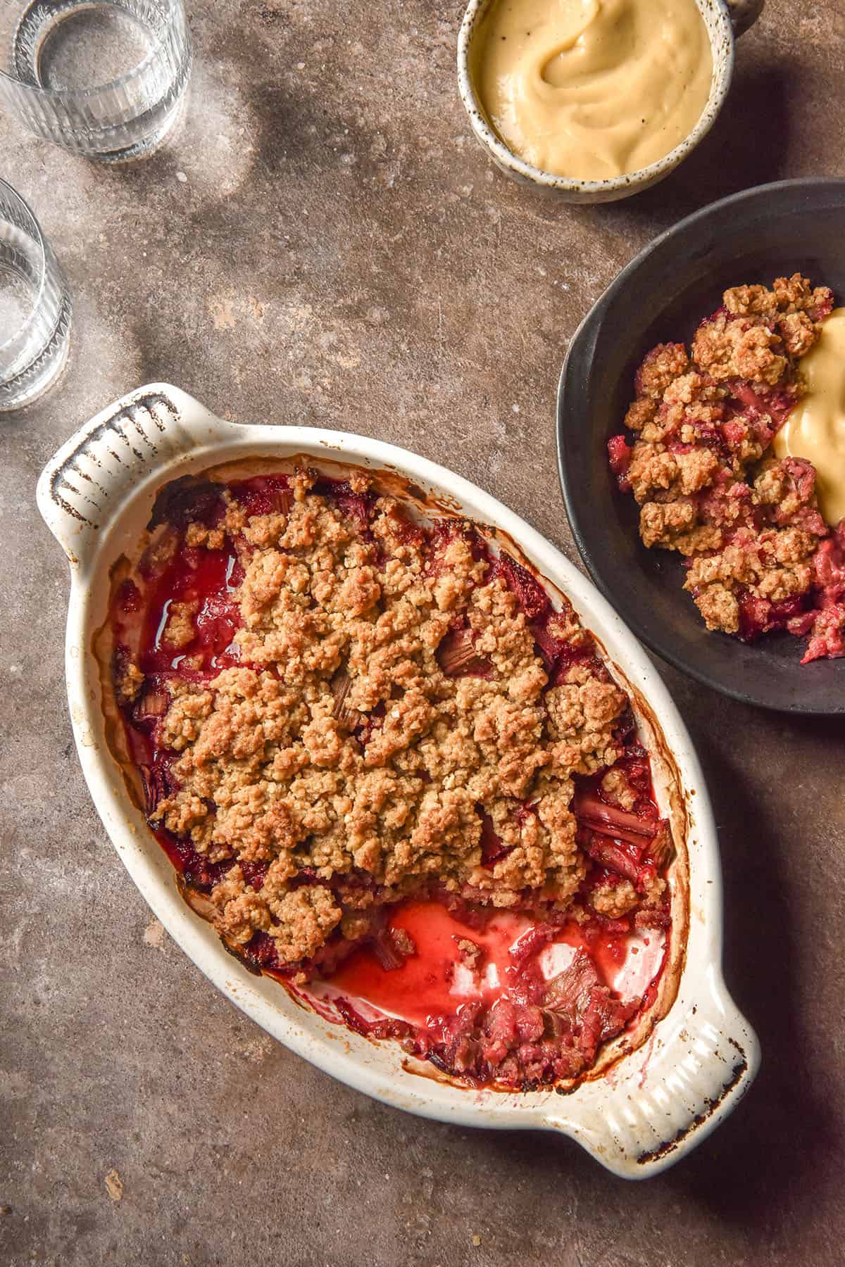 An aerial image of a white oval ceramic dish filled with gluten free rhubarb crumble. The dish sits on a dark brown backdrop and is surrounded by a water glass, a ceramic dish filled with dairy free custard and a bowl filled with crumble and custard