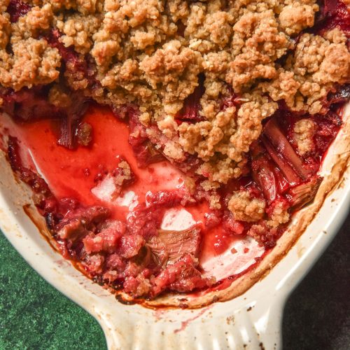 A close up aerial image of a white oval baking dish filled with gluten free rhubarb crumble. The dish sits atop an olive green backdrop