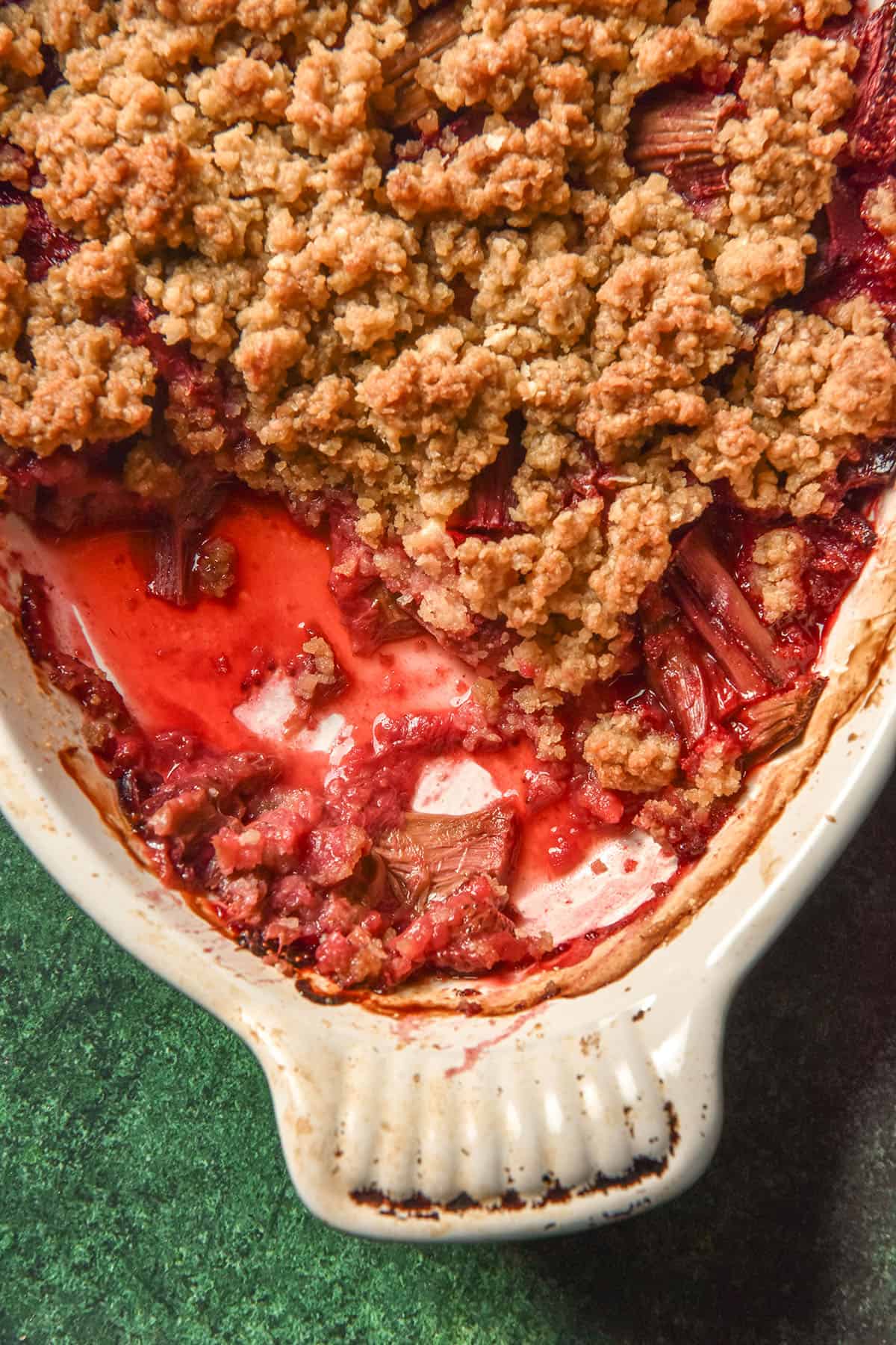 A close up aerial image of a white oval baking dish filled with gluten free rhubarb and strawberry crumble. The dish sits atop an olive green backdrop