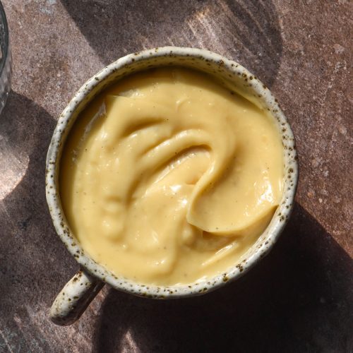 An aerial image of a white ceramic cup filled with lactose free custard. The cup sits on a grey backdrop in contrasting sunlight and the shadows of two glasses of water surround the cup of custard.