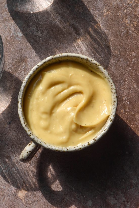 An aerial image of a white ceramic cup filled with lactose free custard. The cup sits on a grey backdrop in contrasting sunlight and the shadows of two glasses of water surround the cup of custard.