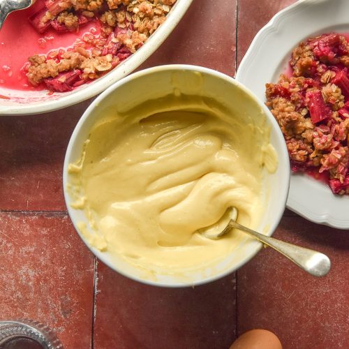 An aerial image of a bowl of lactose free custard on a terracotta tile backdrop. The custard is surrounded by a gluten free rhubarb crumble, eggs, a plate of crumble and glasses of water.