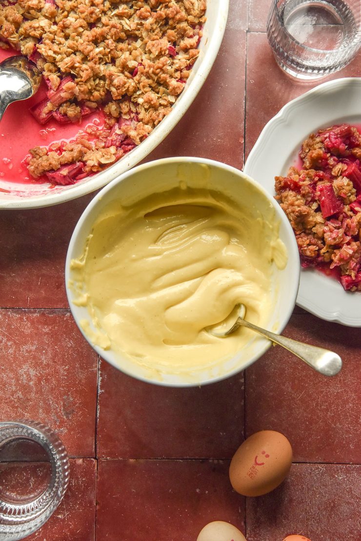 An aerial image of a bowl of lactose free custard on a terracotta tile backdrop. The custard is surrounded by a gluten free rhubarb crumble, eggs, a plate of crumble and glasses of water.