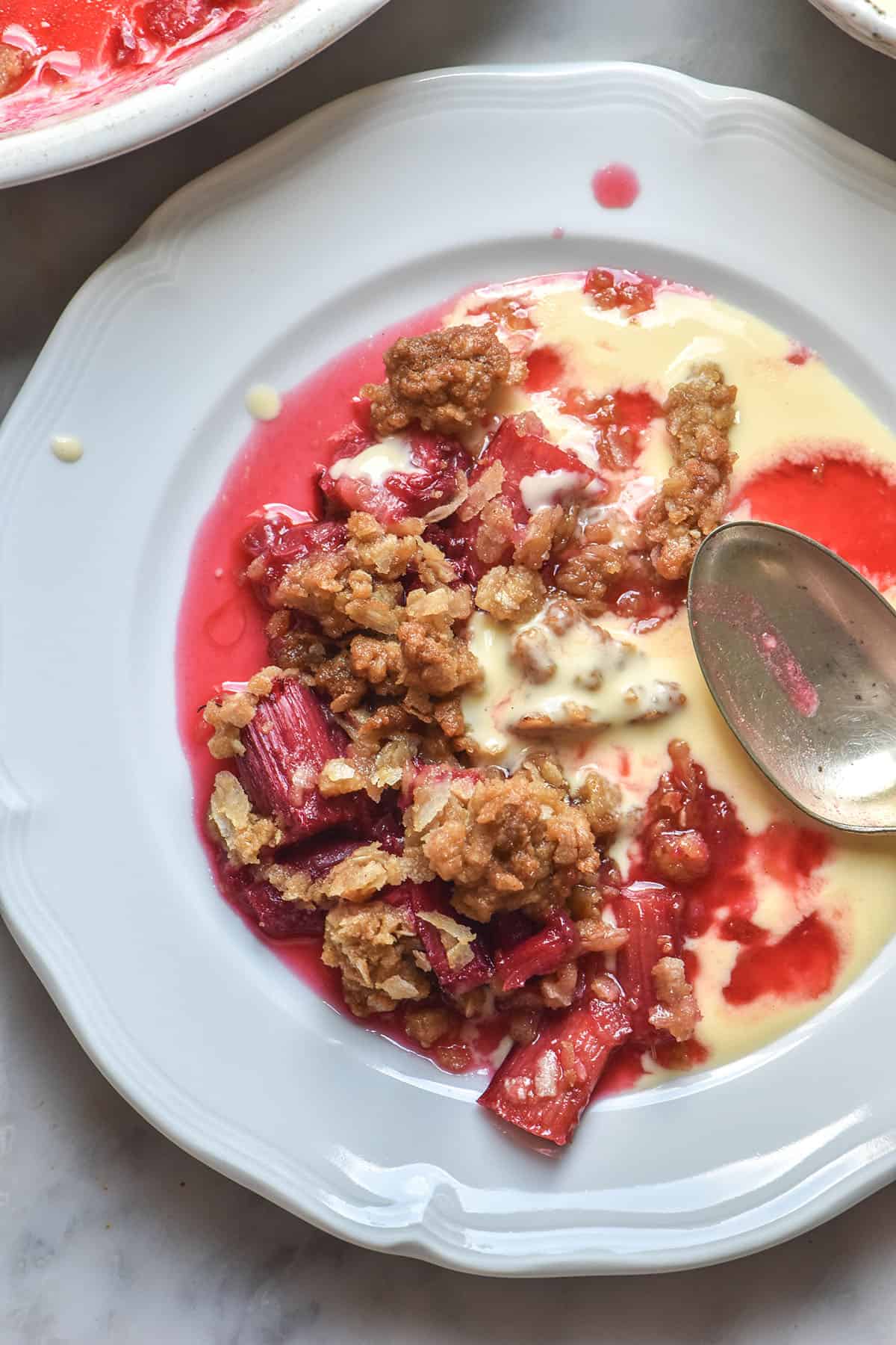 An aerial image of a white scalloped plate topped with gluten free rhubarb crumble and lactose free custard. 