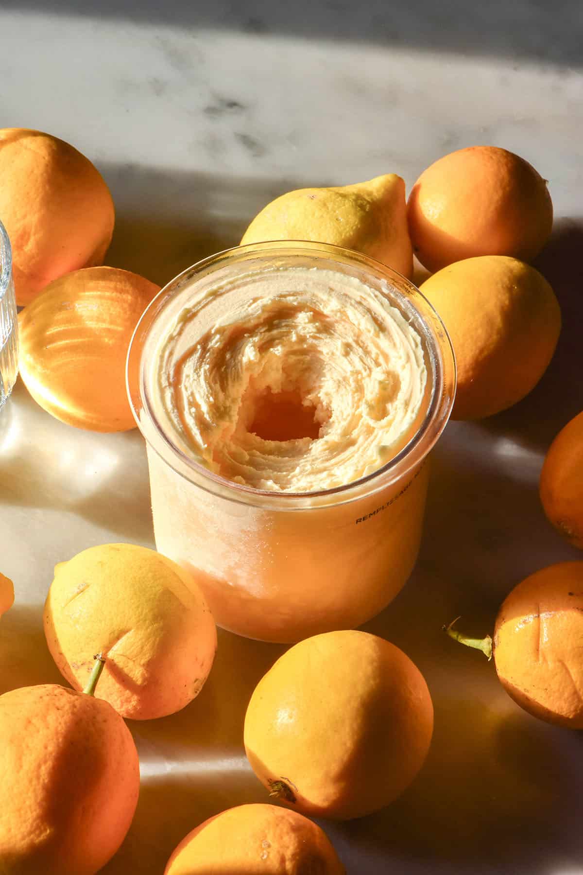 A tub of Ninja Creami lemon curd ice cream sits on a white marble table in contrasting sunlight. The tub is surrounded by Meyer lemons and sunlit glasses of water sit to the left of the image