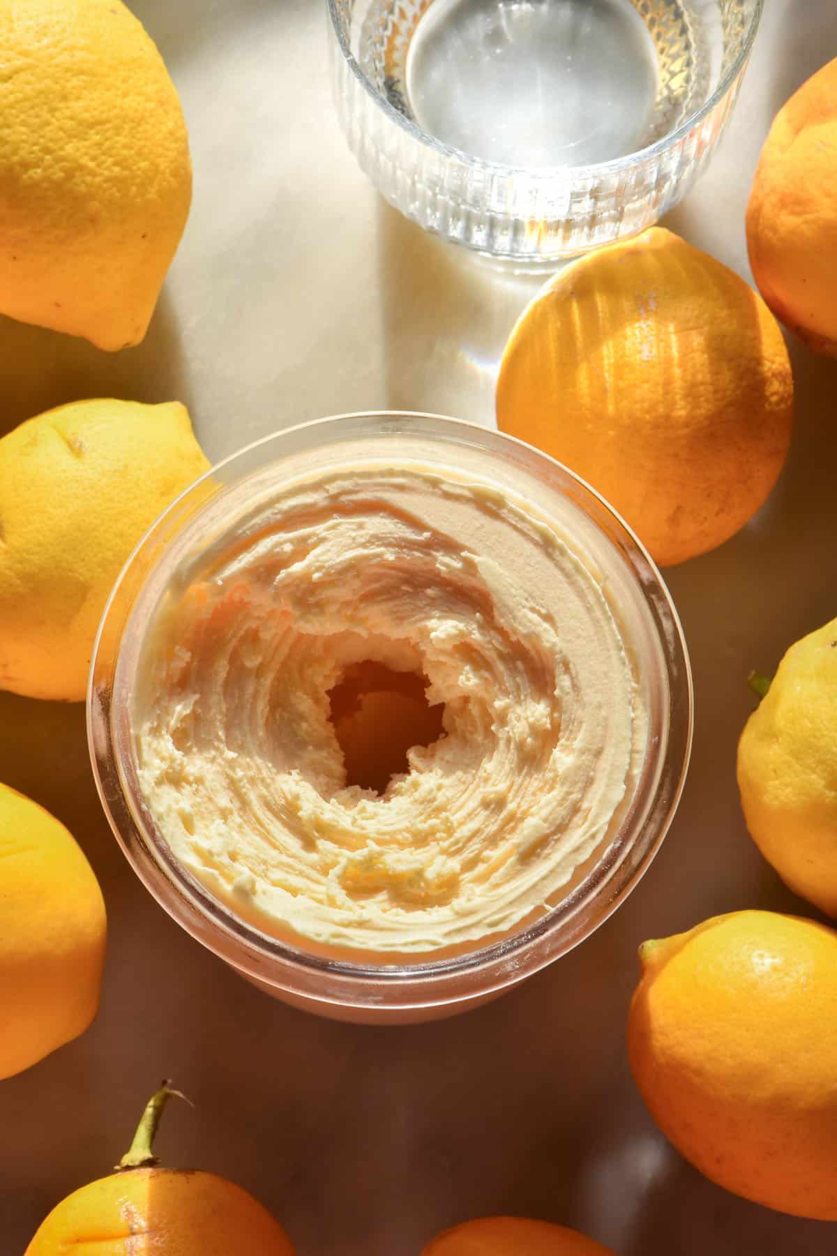 An aerial image of a freshly spun tub of Ninja Creami dairy free lemon ice cream. The tub sits on a sunlit white marble table surrounded by lemons and a sunlit glass of water