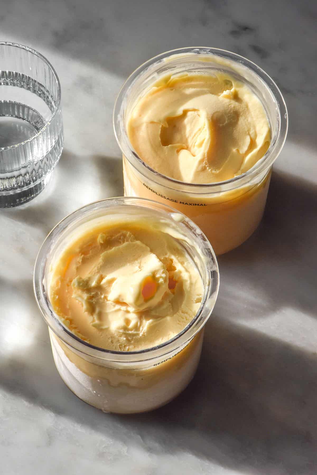 A side on aerial image of two tubs of Ninja Creami lemon ice cream on a white marble table in contrasting sunlight. 