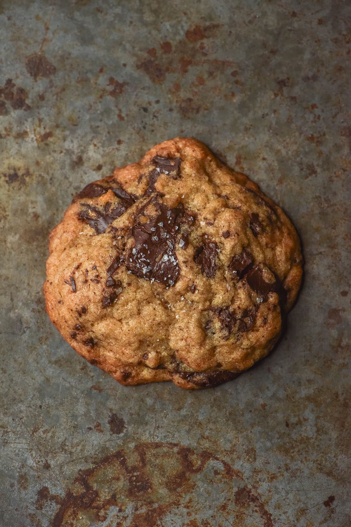 An aerial image of a gluten free banana choc chip cookie on a dark steel backdrop. The cookie is topped with sea salt flakes 