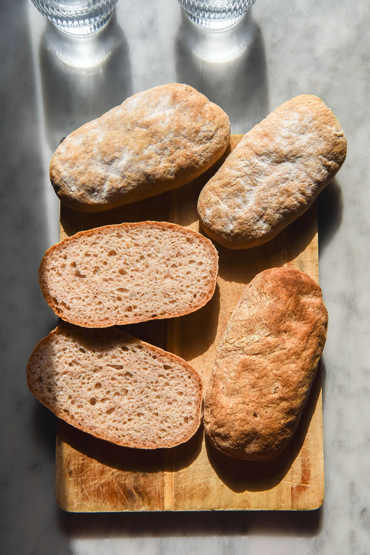 An aerial image of gluten free ciabatta rolls on a wooden board atop a white marble table in bright sunlight