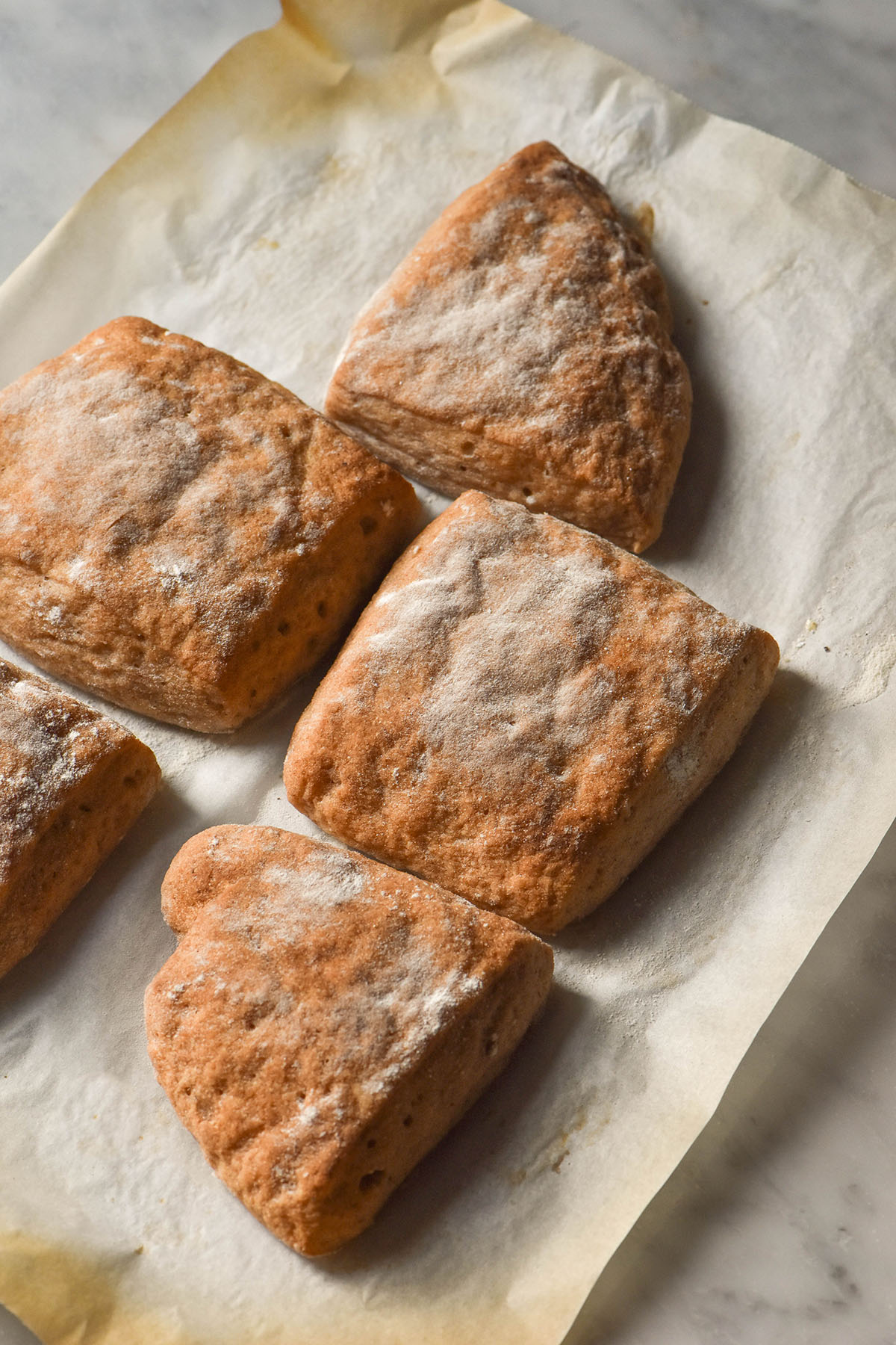 A side on image of gluten free ciabatta rolls on a sheet of baking paper atop a white marble bench top