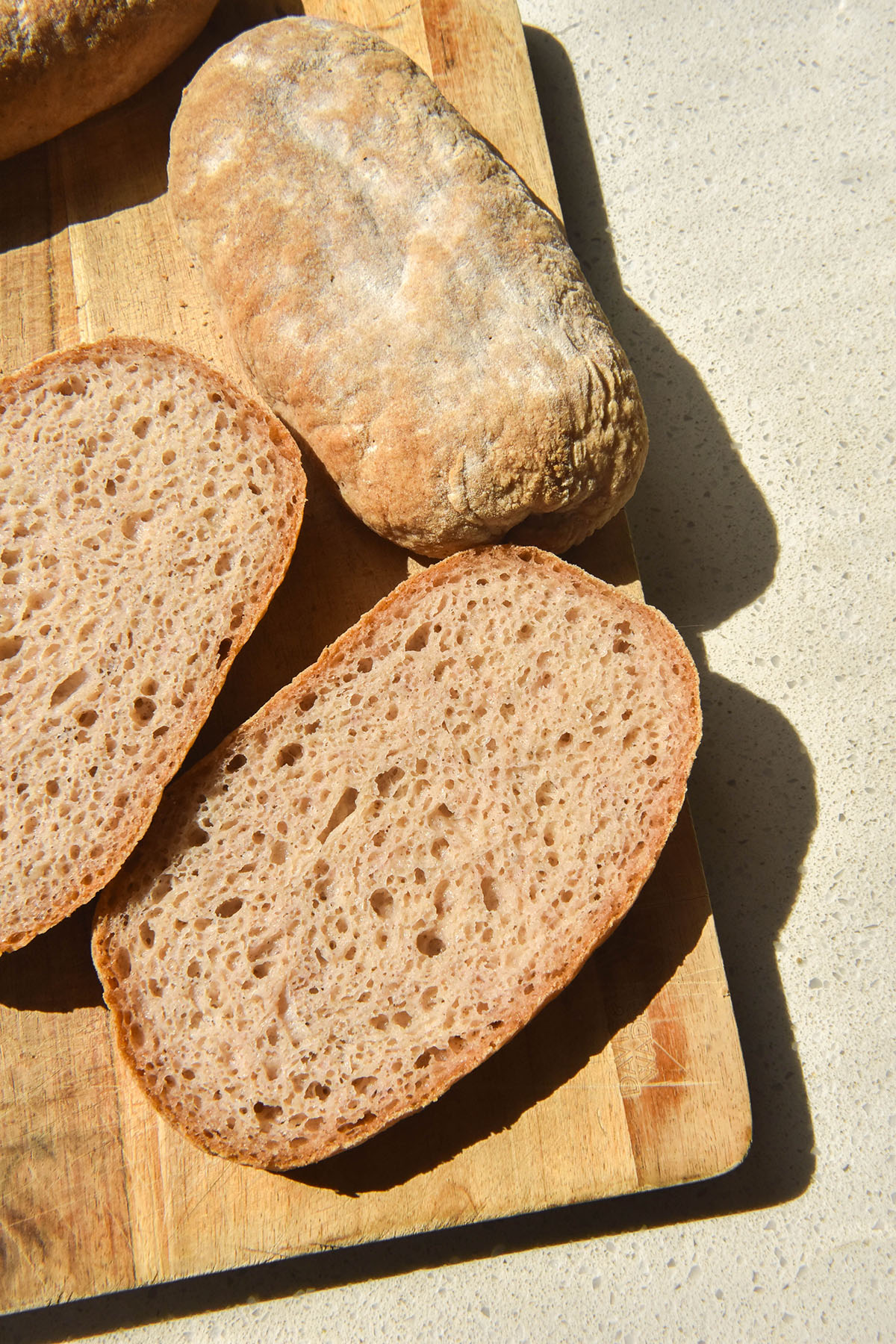 A brightly lit aerial image of gluten free ciabatta buns on a wooden board atop a white stone bench top