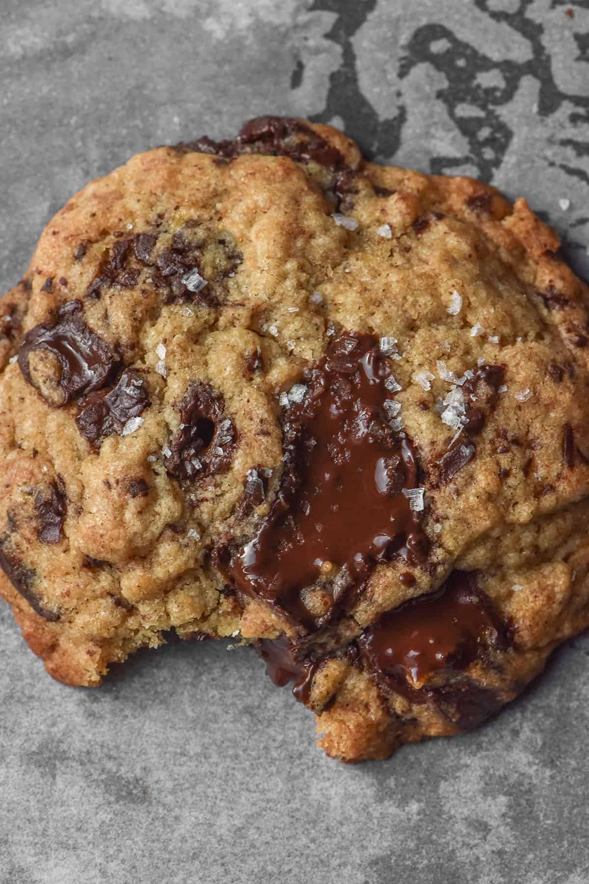 A close up aerial macro image of a gluten free banana choc chip cookie on a lined baking tray. The cookie has a bit taken out of it and a sprinkling of sea salt flakes on top.