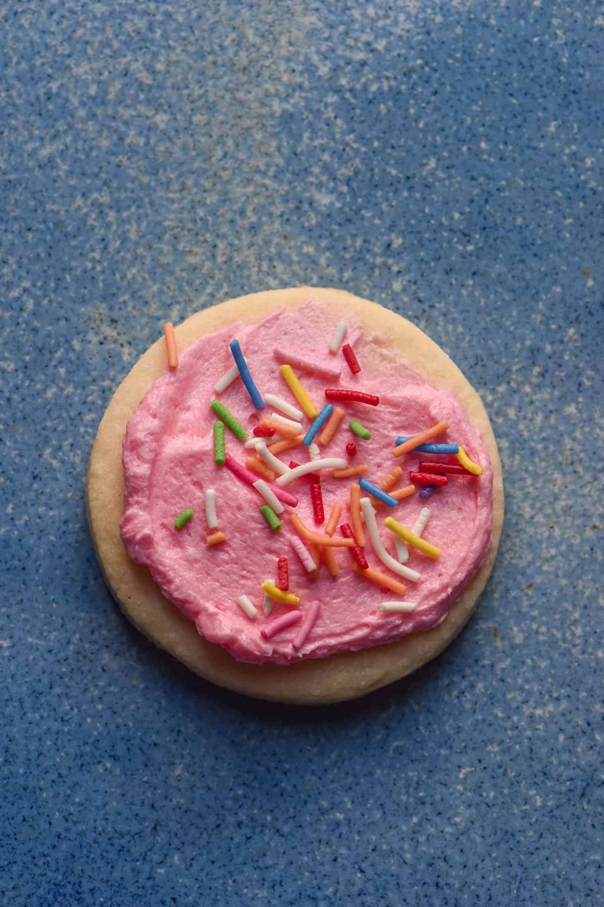 An aerial image of a gluten free sugar cookie topped with pink icing and rainbow sprinkles on a blue ceramic plate.