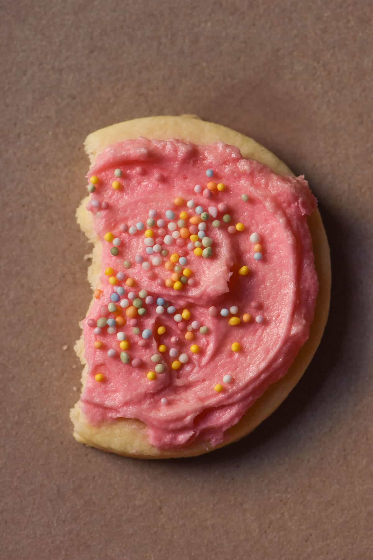 An aerial image of a gluten free sugar cookie topped with pink icing and rainbow sprinkles. The cookie sits on a pale pink ceramic plate and has a bite taken out of the left side of the cookie. 