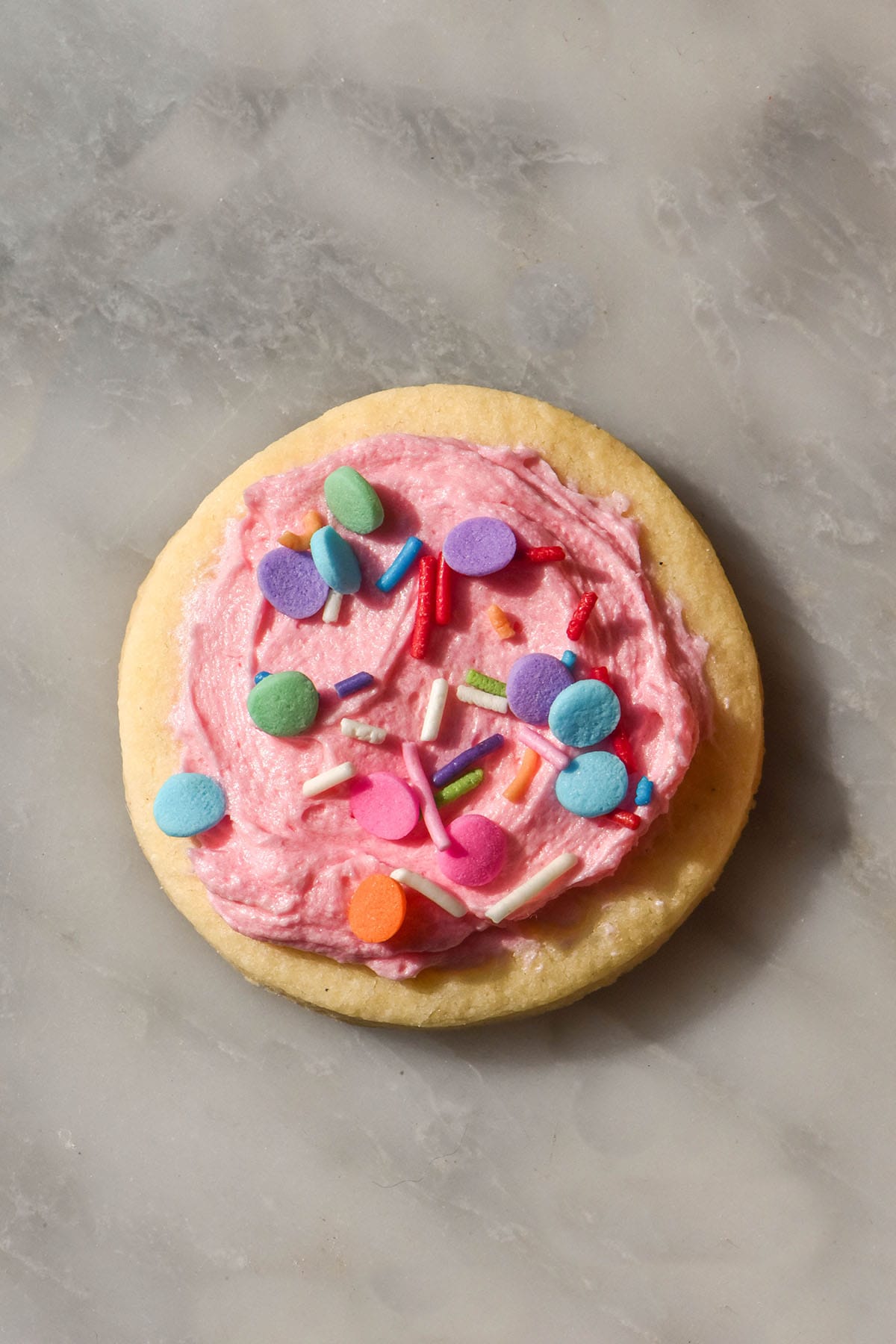 An aerial image of a gluten free sugar cookie on a white marble table. The cookie is topped with pink icing and rainbow sprinkles.