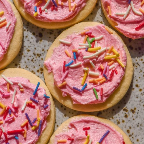 An aerial sunlit image of gluten free sugar cookies on a white speckled ceramic plate topped with pink buttercream and rainbow sprinkles