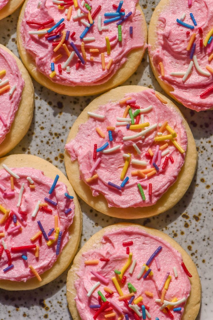 An aerial sunlit image of gluten free sugar cookies on a white speckled ceramic plate topped with pink buttercream and rainbow sprinkles