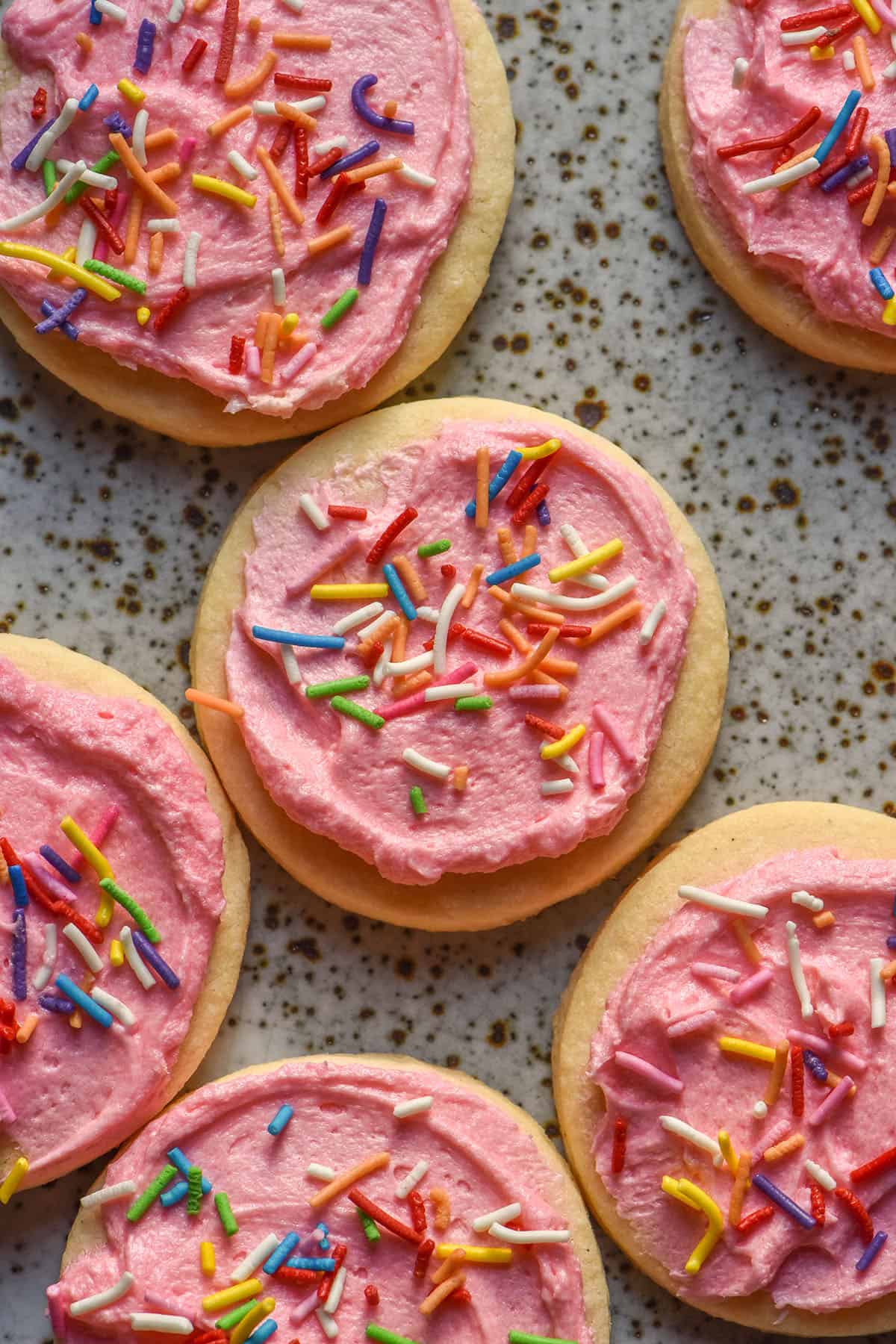 An aerial sunlit image of gluten free sugar cookies on a white speckled ceramic plate topped with pink buttercream and rainbow sprinkles