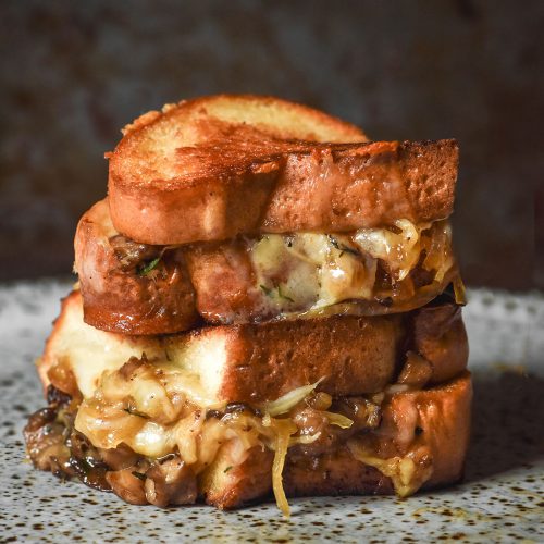 An image of a Low FODMAP caramelised onion and mushroom grilled cheese that is golden brown, sliced in half and stacked on top of each other. The grilled cheese sits on a white speckled ceramic plate against a dark backdrop