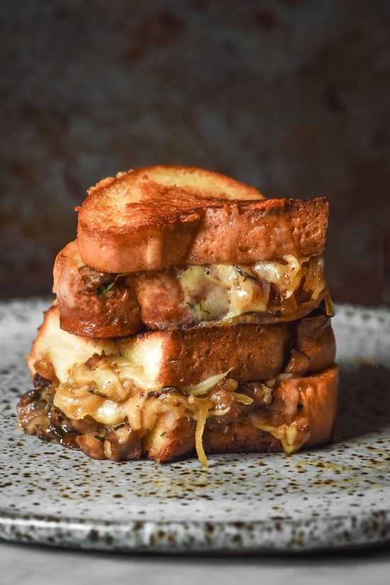 An image of a Low FODMAP caramelised onion and mushroom grilled cheese that is golden brown, sliced in half and stacked on top of each other. The grilled cheese sits on a white speckled ceramic plate against a dark backdrop