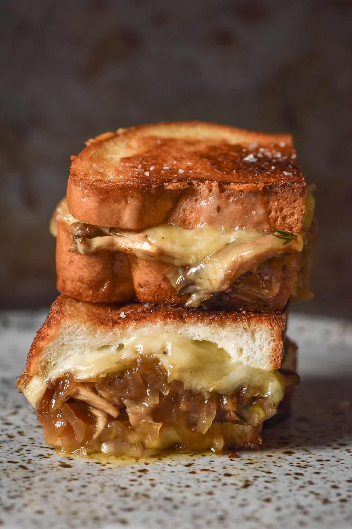 An image of a Low FODMAP caramelised onion and mushroom grilled cheese that is golden brown, sliced in half and stacked on top of each other. The grilled cheese sits on a white speckled ceramic plate against a dark backdrop