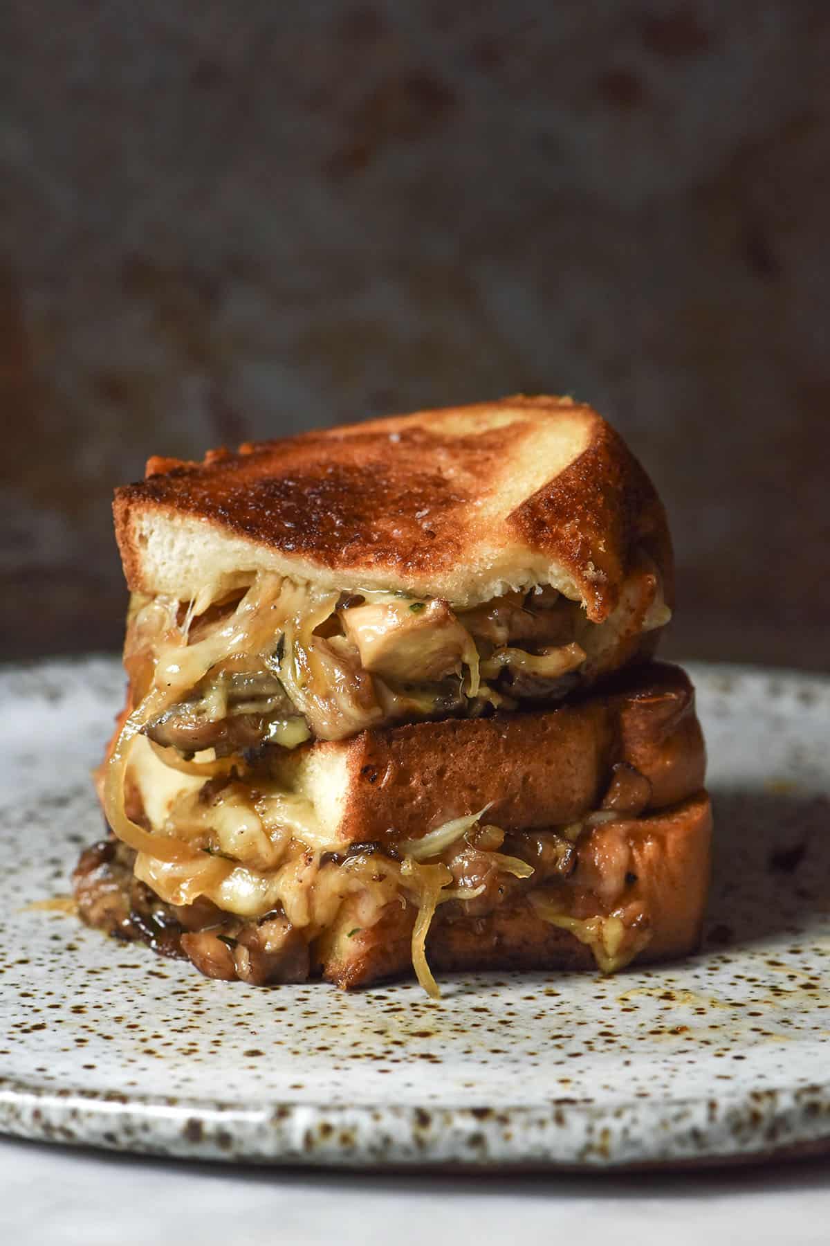 An image of a Low FODMAP caramelised onion and mushroom grilled cheese that is golden brown, sliced in half and stacked on top of each other. The grilled cheese sits on a white speckled ceramic plate against a dark backdrop