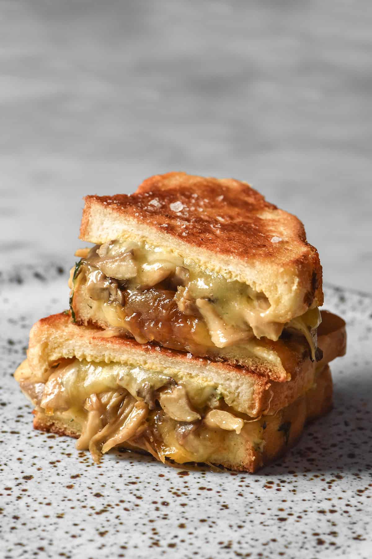 A side on image of a gluten free caramelised onion and mushroom toastie that has been sliced in half and stacked on a white speckled ceramic plate atop a white marble table