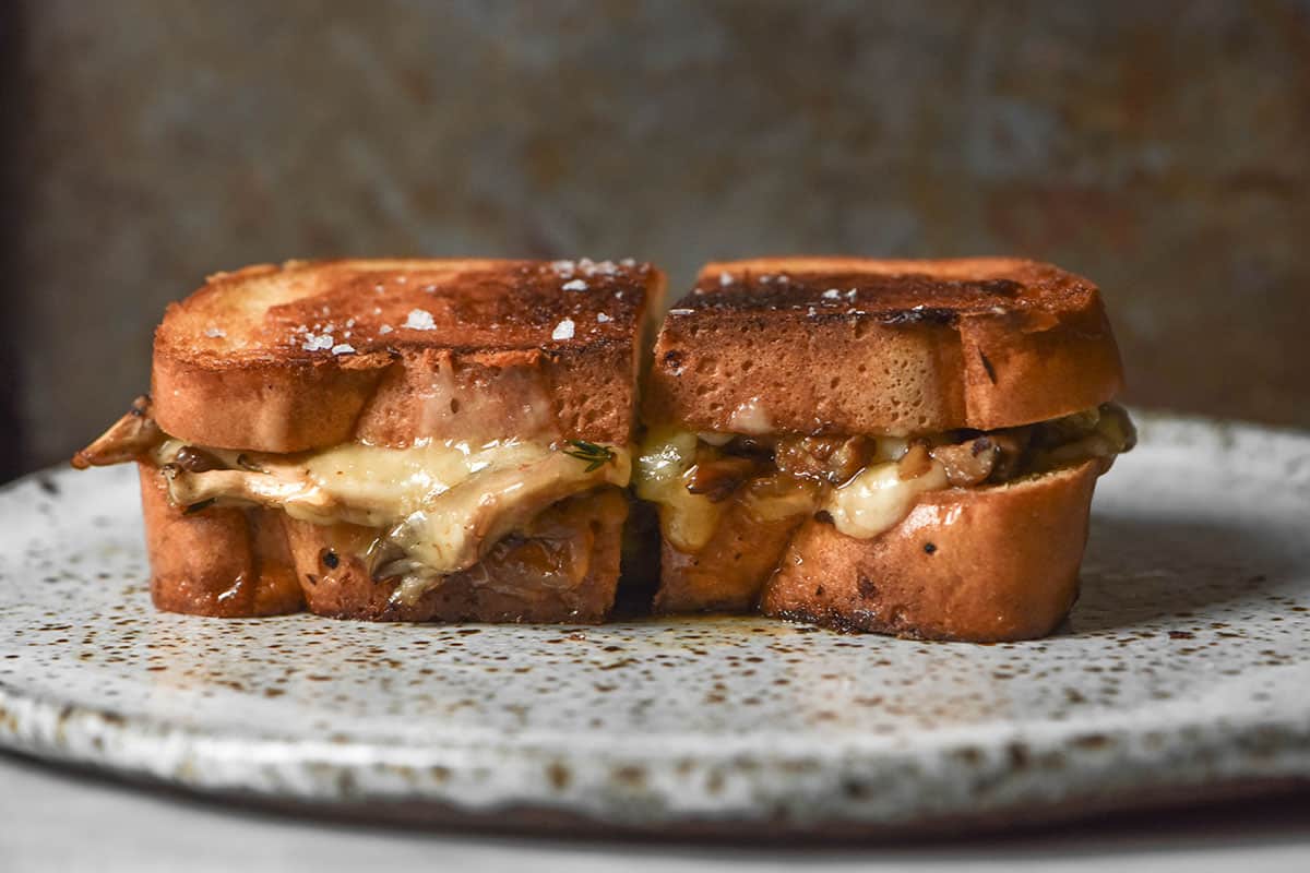 A side on image of a gluten free grilled cheese filled with mushrooms and caramelised onion. The sandwich sits on a white speckled ceramic plate against a dark backdrop