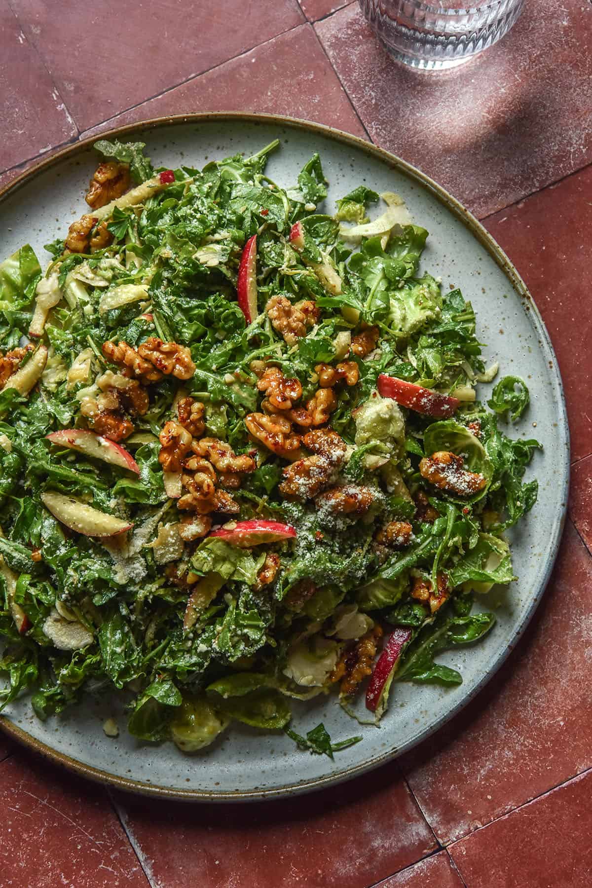 An aerial image of a large white ceramic plate topped with a lower FODMAP shaved Brussels sprout salad with rocket, red apple, maple chilli walnuts and a dusting of parmesan. The plate sits on a terracotta tile backdrop and a glass of water sits in the top right of the image