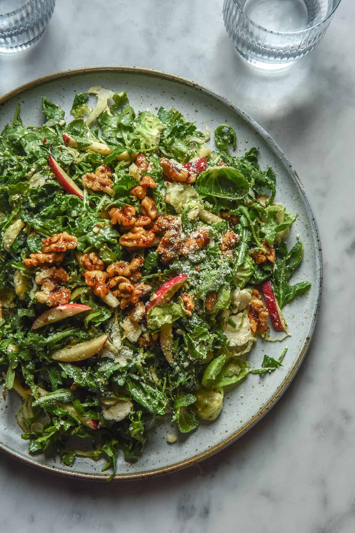 An aerial image of a white ceramic plate topped with a lower FODMAP shaved Brussels sprout salad with rocket, red apple, maple chilli walnuts and a sprinkling of parmesan. The plate sits atop a white marble table and two glasses of water sit above it. 