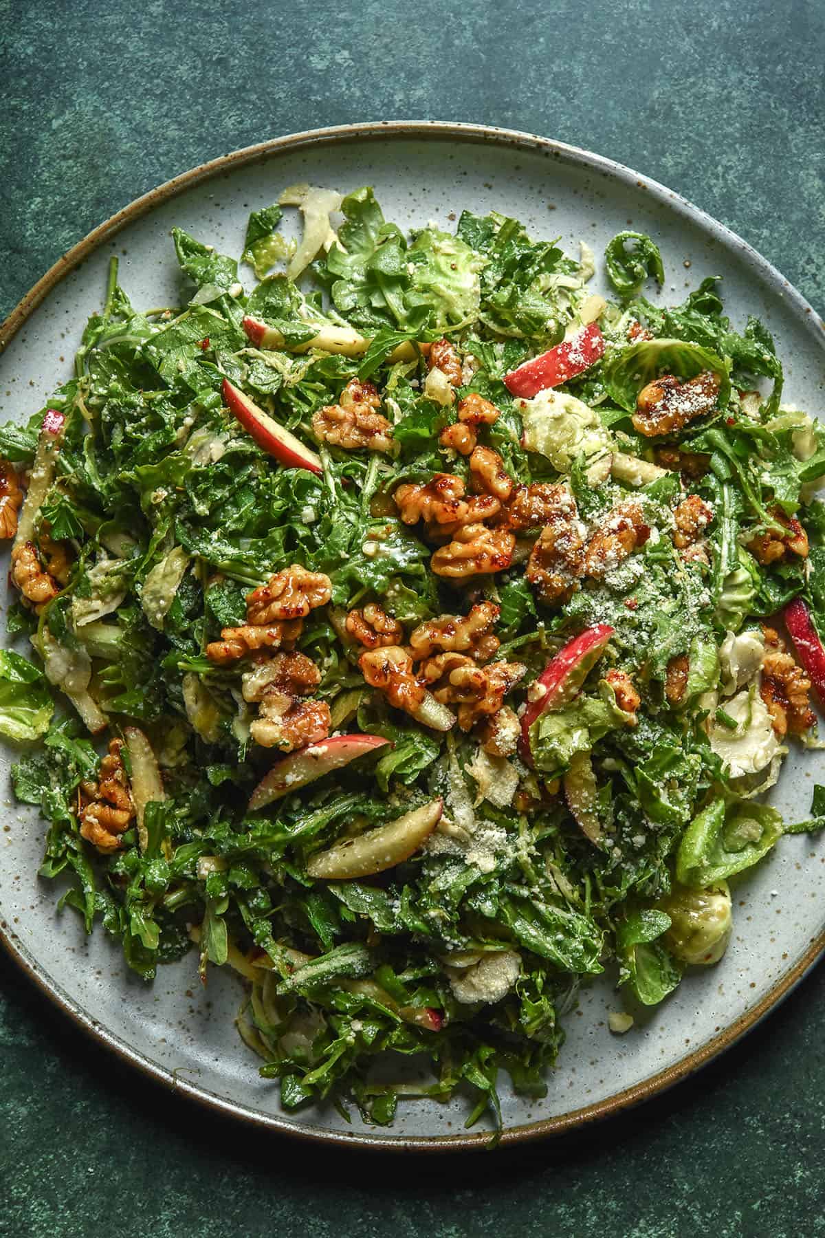 An aerial image of a white ceramic serving plate topped with a lower FODMAP shaved Brussels sprout salad with rocket, red apple, maple chilli walnuts and a sprinkling of parmesan. The plate sits atop a dark olive backdrop