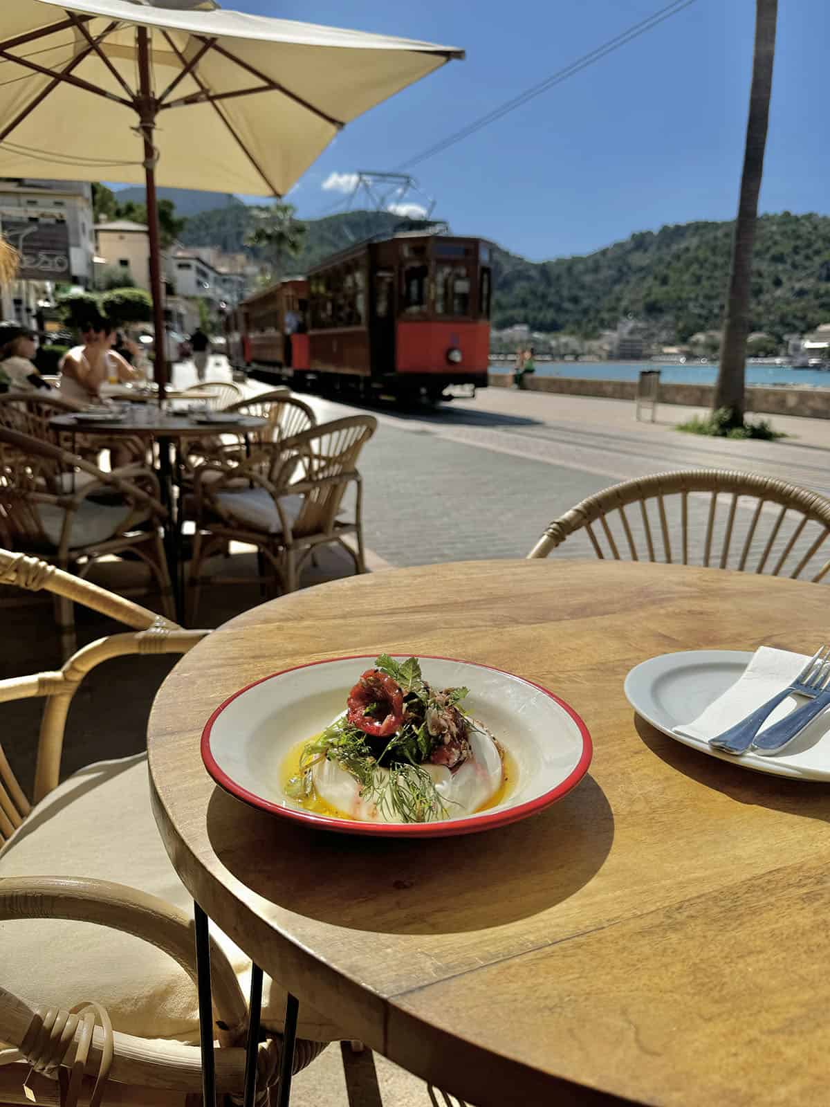 The view from Blai Bar in Port De Soller, Mallorca. A burrata dish sits on the table in the foreground while the Soller tram goes past the ocean in the background