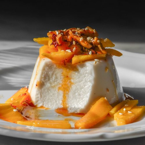 A side on image of a coconut panna cotta topped with mango and chilli crisp on a white plate in bright contrasting sunlight