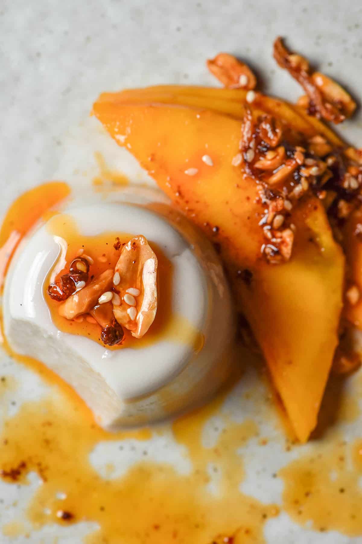 An aerial image of a vegan coconut panna cotta topped with chilli crisp and mango on a white plate atop a white marble table